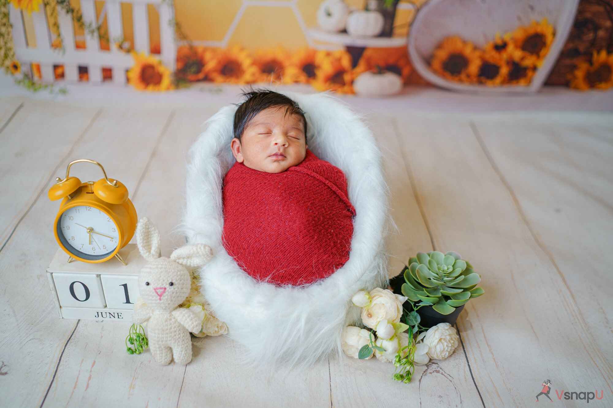 A baby sleeping peacefully in a white fur bed with a red sheet, surrounded by date props and delicate flowers in a heartwarming baby photoshoot.